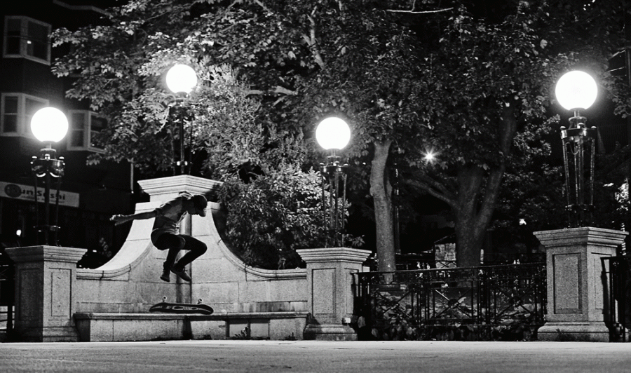 Black and white photo of a Boy doing a skateboard trick off a bench at night in a park.