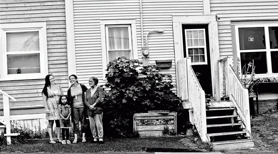 Black and white photo 4 women pose for a photo standing in front of a house smiling