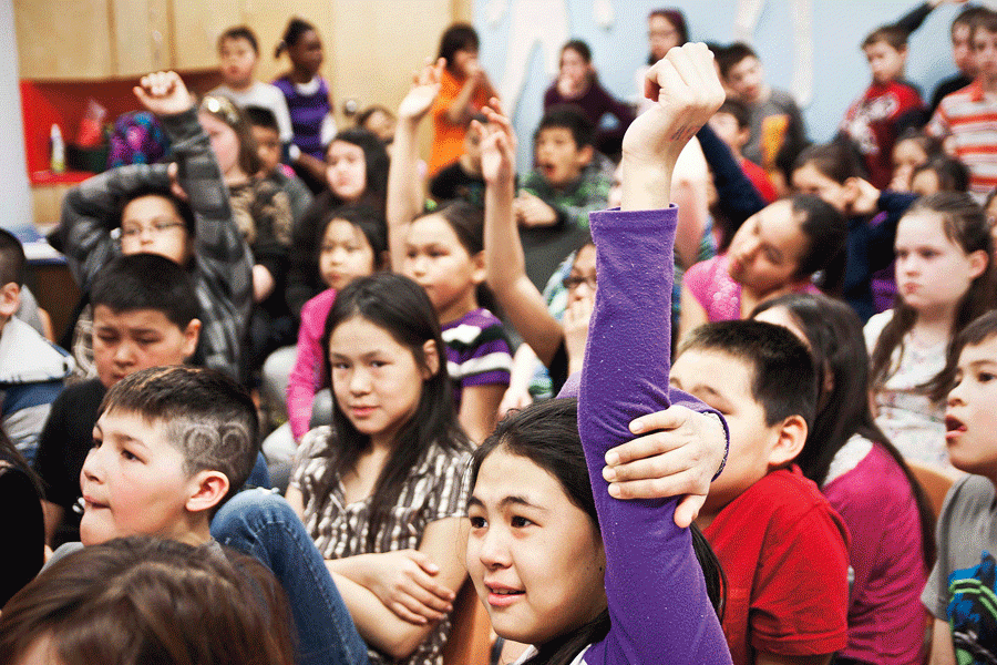 Children sit in a large group and some raise their hand to answer a question